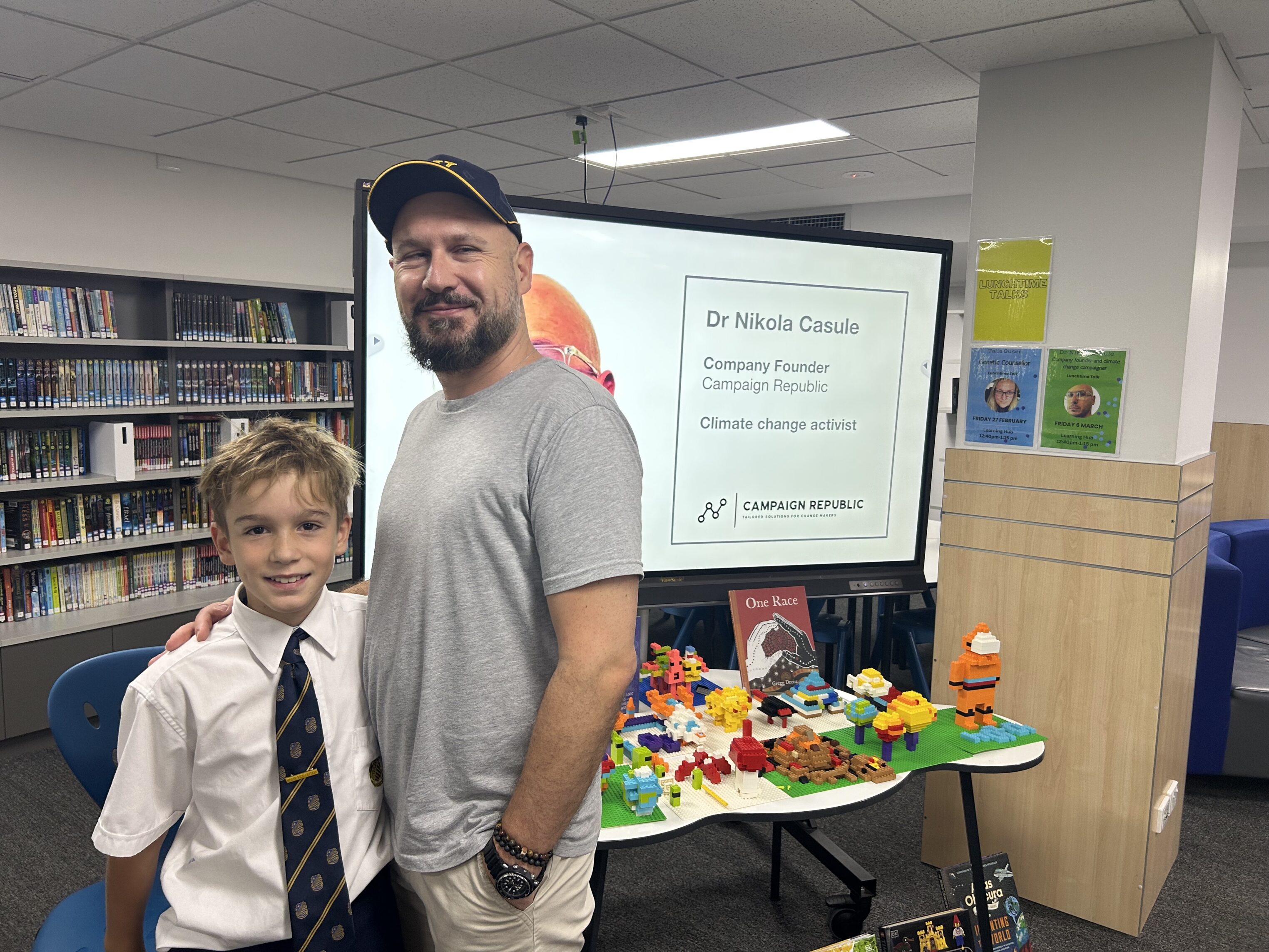A man and a boy stand smiling in a library. Behind them is a screen displaying Dr Nikola Casule, Company Founder, Campaign Republic, Climate change activist. Colourful LEGO models are on a table in front of the screen.