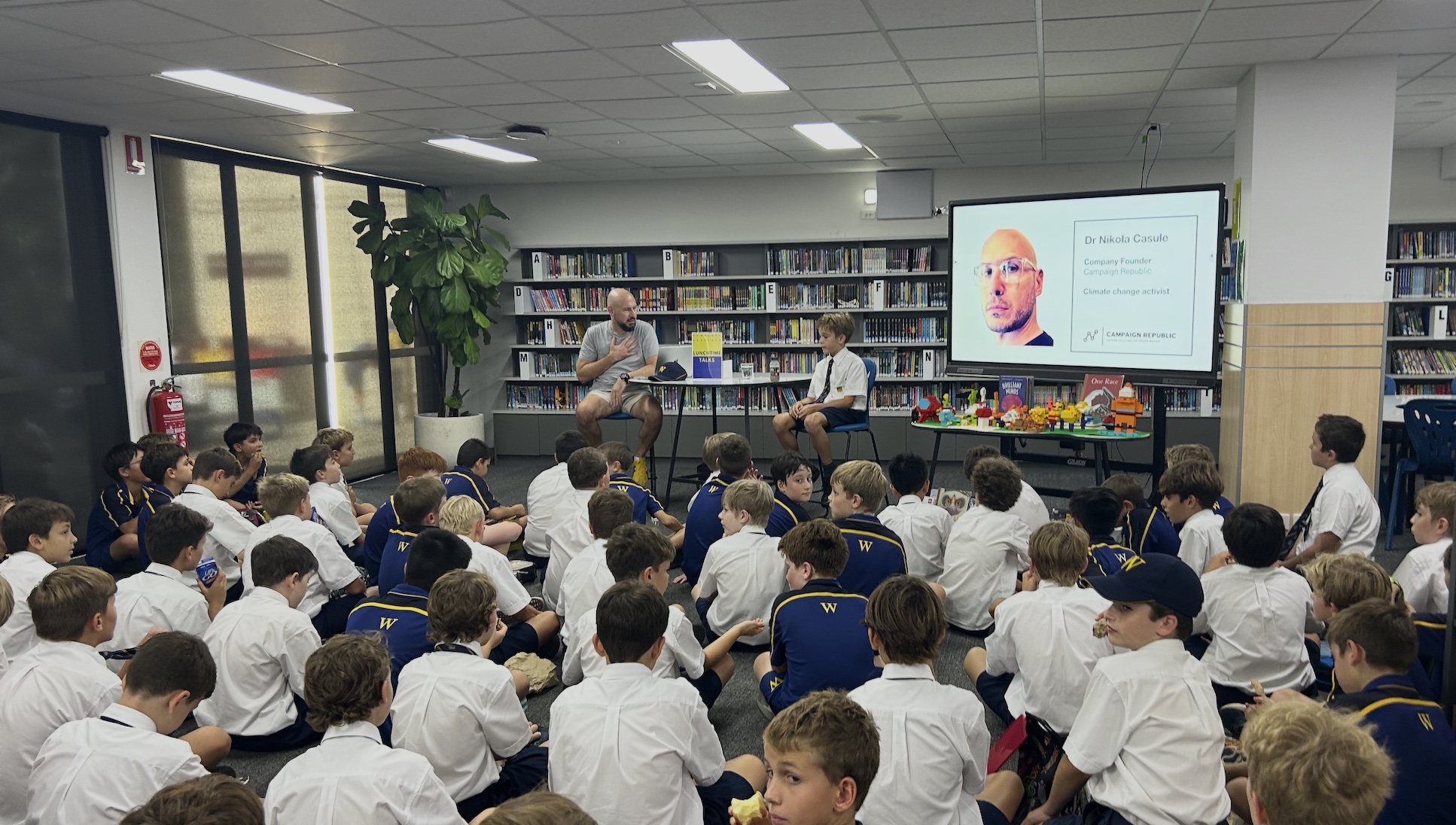 A large group of schoolboys in uniforms sit on the floor of a library, listening to a guest speaker and a student interviewer seated at the front, with a presentation slide of Dr Nichola Conliffe displayed on a screen behind them.