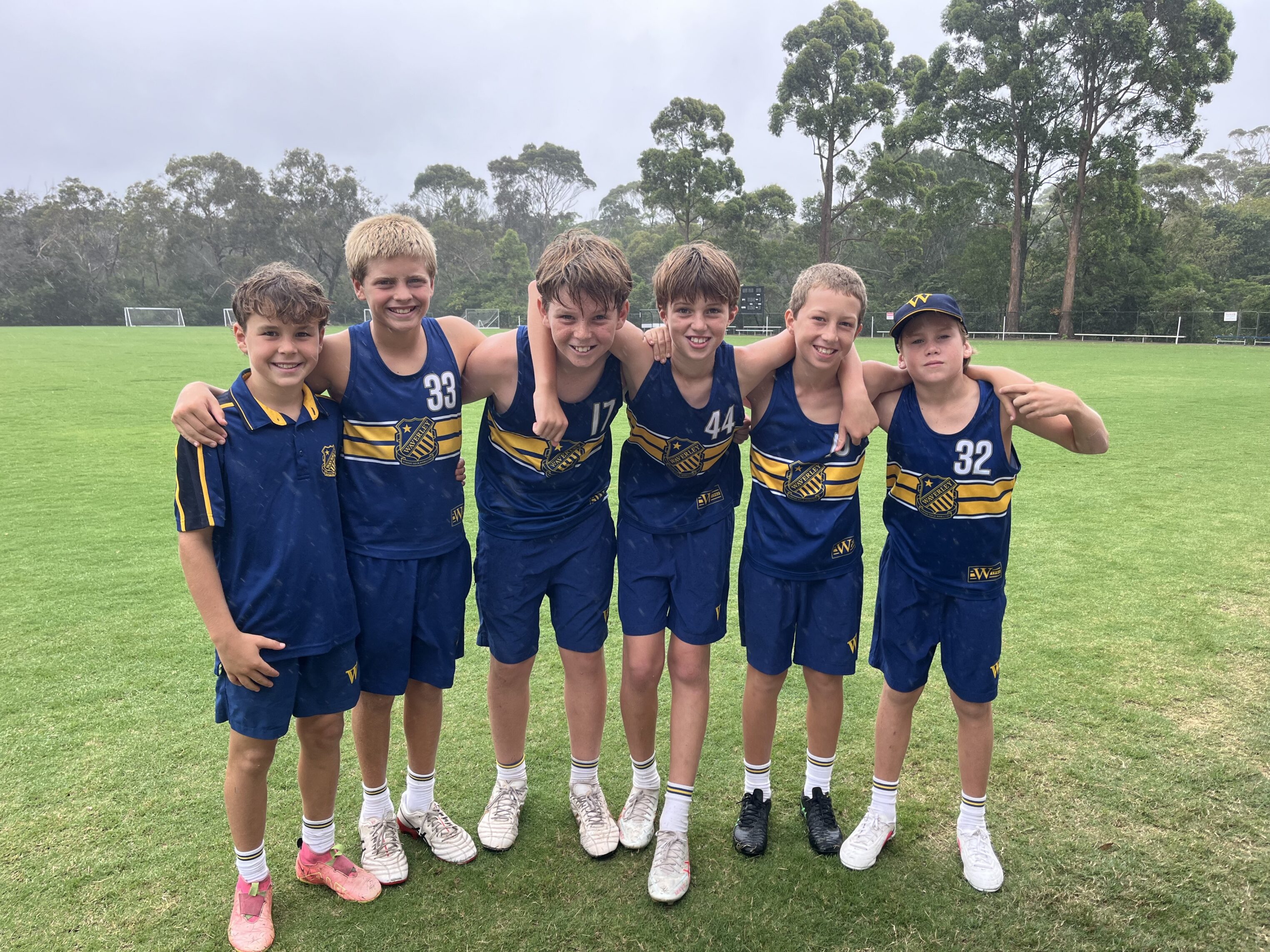 Six boys in blue and yellow sports kit stand side by side on a grassy pitch, smiling and posing with their arms round each other. Trees and goalposts are visible in the background.