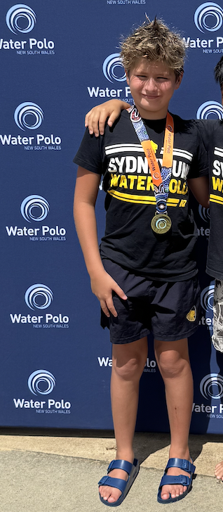 A smiling child wearing a Sydney Uni Water Polo shirt, dark shorts, and blue sandals stands in front of a Water Polo New South Wales backdrop, displaying a medal around their neck.