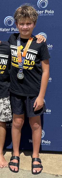 A young boy wearing a black Uni Water Polo t-shirt, shorts, and sandals stands in front of a Water Polo New South Wales backdrop, smiling and wearing two medals round his neck.