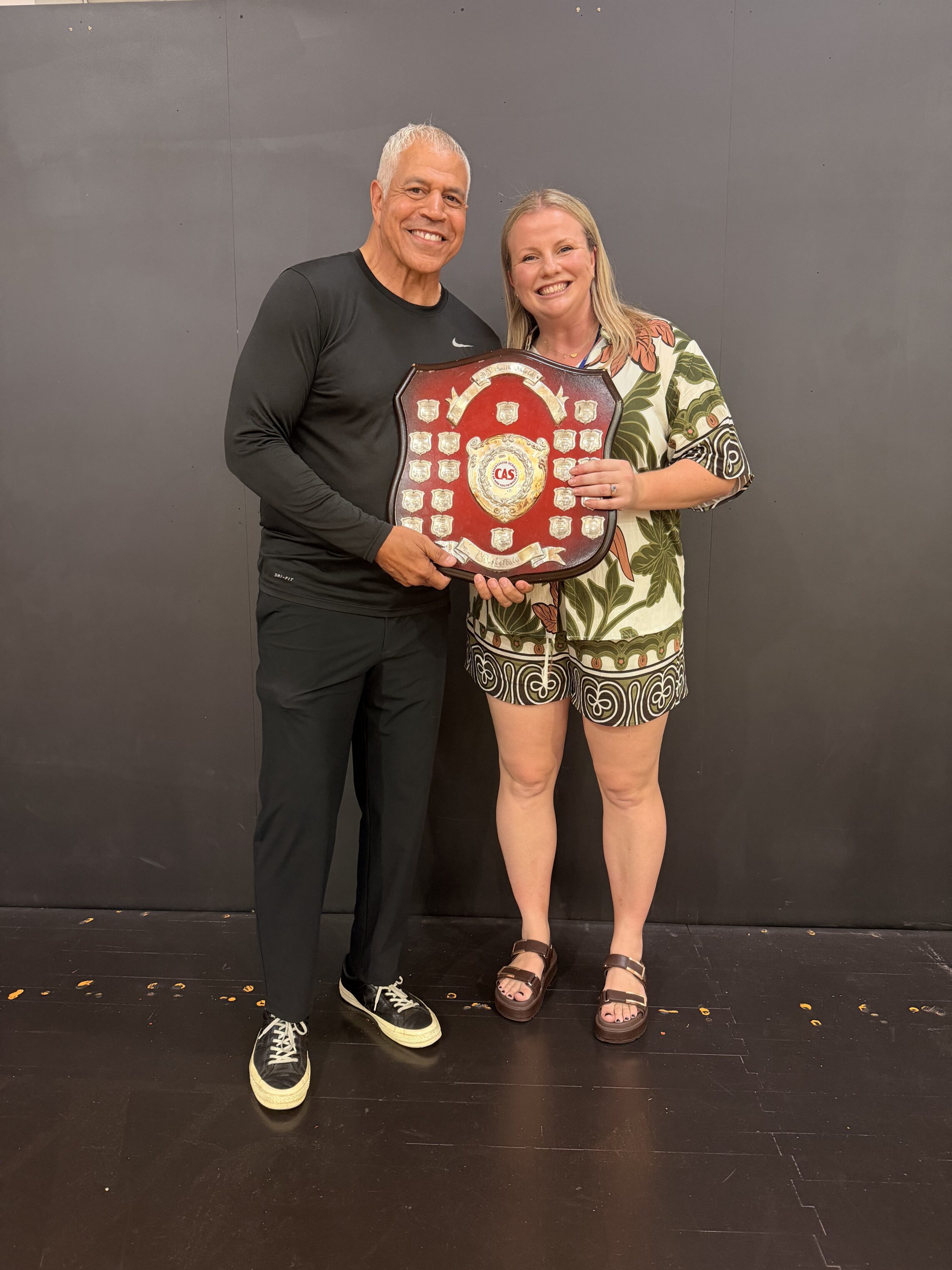 Two people stand smiling on a dark stage, holding a large red and silver award plaque together. The man is dressed in black and the woman in patterned shorts and a top.