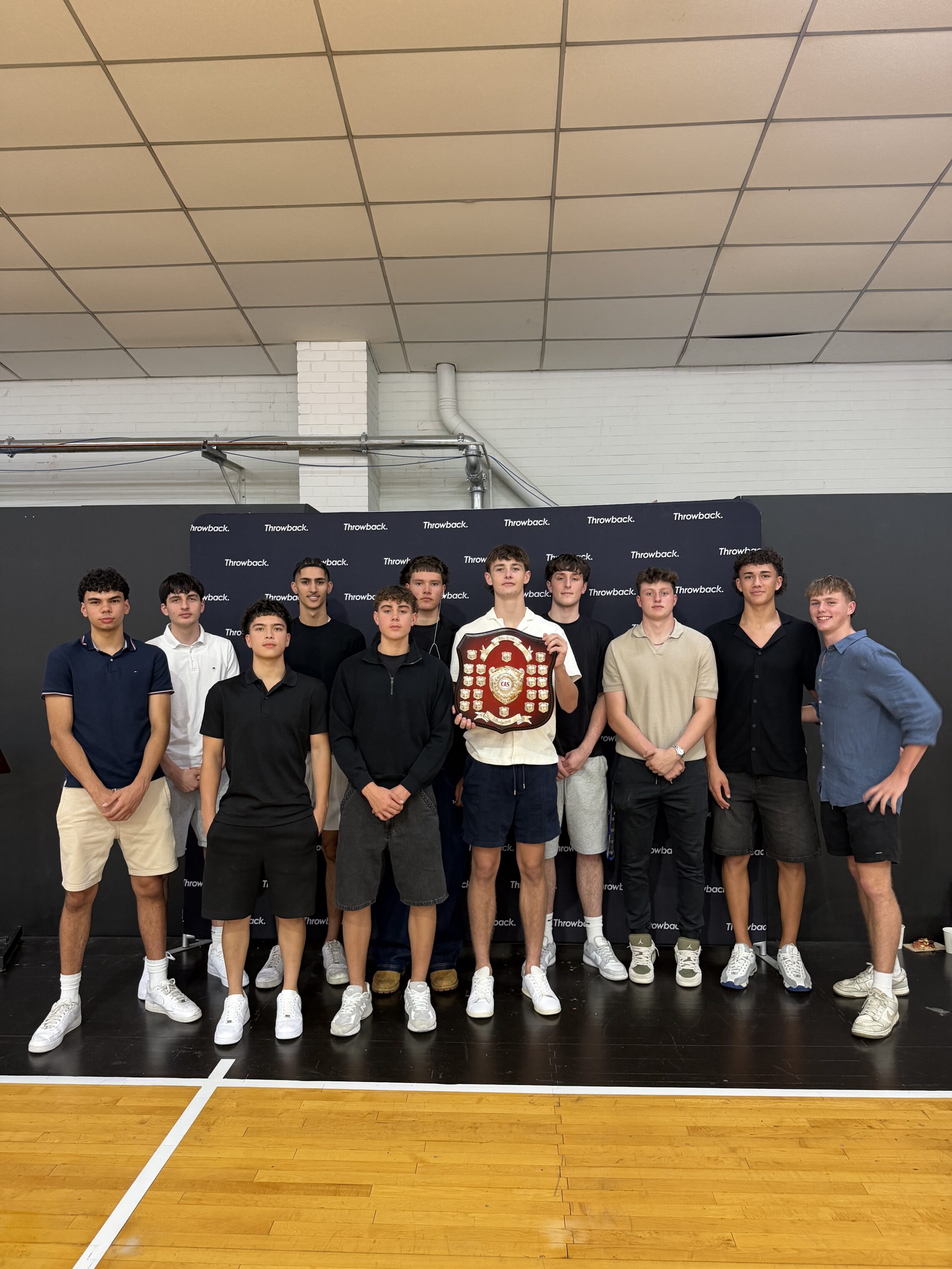 A group of 13 young men pose indoors on a sports court, with one person in the centre holding a shield-shaped trophy. A Powerdeck-branded backdrop is behind them. Most are wearing casual outfits and white trainers.