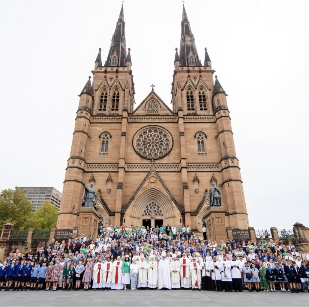 A large group of people, including clergy in white robes and others in uniforms, stands on the steps in front of a grand cathedral with twin spires and a large rose window.