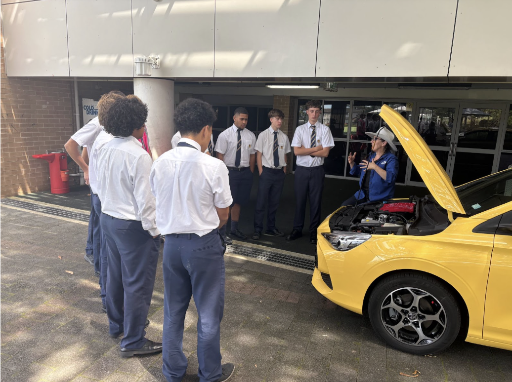 A group of students in school uniforms stand around a yellow car with its bonnet open, listening to an instructor who is explaining something about the engine in an outdoor area near a building.